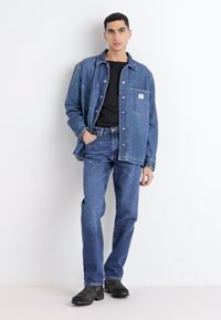Keskmise sinise denimpusa ja teksade komplekt, millel on rinnakott ja nööbid. All on must T-särk, millele on lisatud mustad saapad.