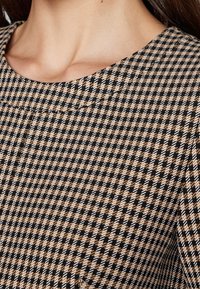 Femme portant un haut beige, noir et blanc à motif pied-de-poule avec un col rond, vue en gros plan.