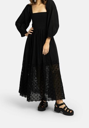 Vrouw draagt een zwarte maxi-jurk met lange mouwen, een gerimpeld lijfje en een laser-gesneden bloemenzoom, gecombineerd met zwarte platformsandalen.