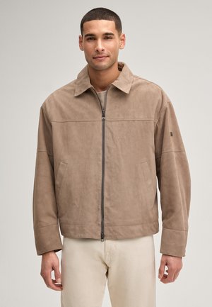 Junger Mann, der eine beige Reißverschluss-Suedejacke mit Kragen und cremefarbene Hosen trägt, steht vor einem schlichten hellen Hintergrund.