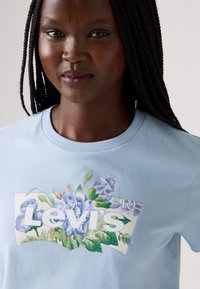 Lyseblå bomull T-skjorte med et blomsterdesign i blå og grønne nyanser, med "Levi's"-tekst i fet hvit skrift i midten.