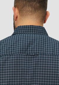 Marineblauwe geblokte shirt met een wit gridpatroon, textuurstof, korte kraag en een lus aan de achterkant. Effen achtergrond.