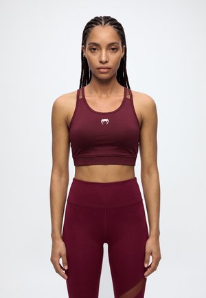 Une femme aux cheveux tressés porte un soutien-gorge de sport bordeaux et un legging assorti sur un fond clair uni.
