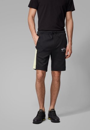 Mand iført sorte shorts med lyse sidepaneler, sort T-shirt, sorte sneakers, stående foran en ensfarvet grå baggrund.