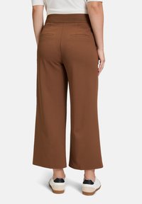 Pantaloni ampi marroni con una vita a coste, due tasche posteriori e una texture liscia, abbinati a sneakers bianche con un accento nero.
