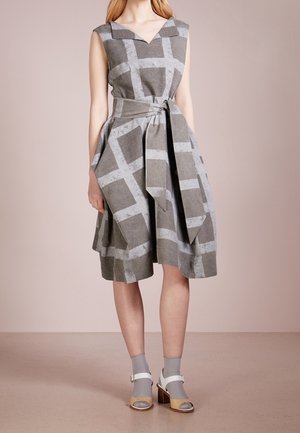 Robe grise sans manches avec un large motif quadrillé gris clair, décolleté en V, taille nouée, portée avec des chaussettes grises et des sandales blanches à talon bloc ouvertes au niveau des orteils.