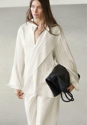 Frau in weißer, strukturierter Jacke und Hose, die eine große schwarze geflochtene Handtasche hält, vor einem schlichten hellen Hintergrund stehend.