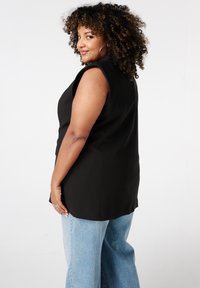 Vrouw met krullend haar, die oorbellen met een ring draagt, een mouwloze zwarte top en lichtblauwe jeans aan heeft, zijwaarts staat en naar de camera glimlacht.