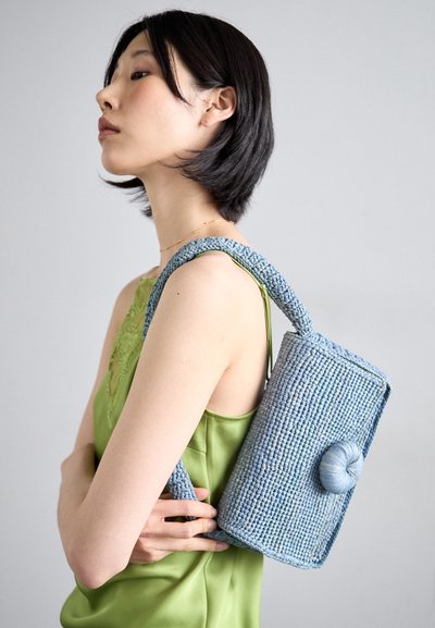 Blue woven handbag with a rounded closure, featuring a textured handle. Model wears a green sleeveless top, showcasing the bag's design.