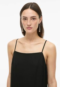 Femme aux cheveux bruns raides portant un haut noir à fines bretelles et de longues boucles d'oreilles argentées en forme de petites boules, sur un fond blanc uni.