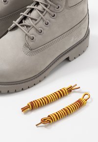 Botte en daim gris à lacets avec semelle épaisse, à côté de deux lacets enroulés jaunes et bruns sur fond blanc.