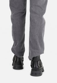 Botas de tobillo de cuero negro con detalles de hebilla, suelas gruesas y pantalones de tela gris texturizada. Enfoque en un diseño funcional y resistente.