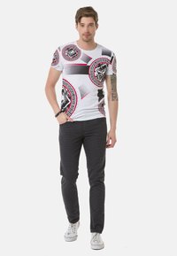 Weißes Grafik-T-Shirt mit kreisförmigen schwarzen und roten Mustern, kombiniert mit grauen gemusterten Hosen und weißen Sneakern, vor einem einfarbigen Hintergrund stehend.