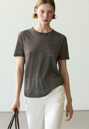 Młoda osoba ubrana w czarno-biały pasiasty krótki rękaw i białe spodnie, trzyma ciemną teksturowaną torbę na jednolitym tle.