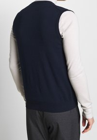 Mörkblå ärmlös tröja med en slät stickad textur, ribbat nederdel och en kontrasterande lättvit långärmad skjorta under.