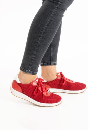 Red mesh sneakers with red lace and accents, white heel cap, and white rubber sole. Worn with dark denim jeans, showing ankle detail.