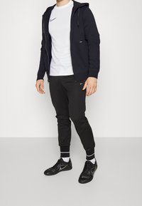 Sweat à capuche zippé bleu marine, t-shirt blanc avec logo, joggers noirs avec logo, chaussures de sport noires ; tenue décontractée de sport.