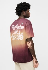 Κοντομάνικο t-shirt με διαβάθμιση χρωμάτων από μοβ σε μπεζ, με μεγάλο λευκό κείμενο "Radiate the" και κυκλικό γραφικό στην πλάτη.