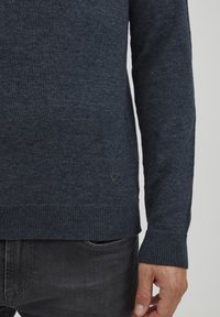 Maglione lavorato a maglia blu scuro con maniche lunghe, polsini e orlo a coste, caratterizzato da un piccolo logo triangolare nero vicino al fondo. Tessuto testurizzato.