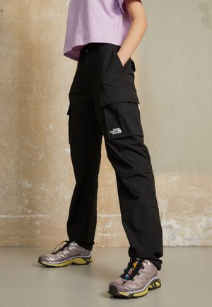 Osoba ubrana w czarne spodnie cargo z logo The North Face i fioletowe buty do biegania w terenie, stojąca na tle beżowej, teksturowanej ściany.
