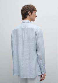 Giovane uomo con i capelli corti, indossa una camicia azzurro chiaro, visto di spalle su uno sfondo neutro, che mostra uno stile informale.