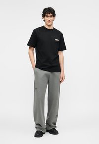 Jeune homme aux cheveux bouclés portant un t-shirt noir imprimé, un pantalon ample gris avec de petits logos, et des baskets noires, debout avec une main dans la poche.