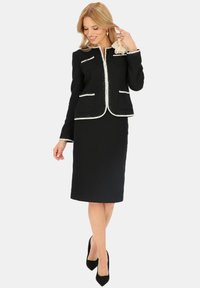 Czarny zestaw w tweedzie składający się z dopasowanej marynarki z białymi wykończeniami, dwoma kieszeniami z przodu oraz spódnicy do kolan. Stylizowany z czarnymi szpilkami.