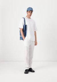 T-shirt bianca in cotone, cappellino da baseball in denim chiaro, pantaloni bianchi larghi, scarpe in pelle nere, borsa a tracolla blu in tela con dettagli testuali.
