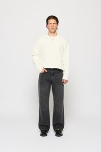 Cremefarvet sweater med V-udskæring, parret med mørkegrå brede jeans. Modellen bærer sorte sko og et guldkæde.