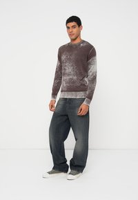 Mann steht und trägt einen braun-grau marmorierten Pullover, lockere dunkelgraue Jeans und grau-weiße Turnschuhe vor einem einfachen weißen Hintergrund.