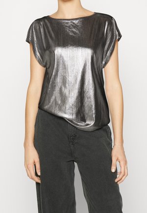 Femme portant un haut métallique brillant à manches courtes associé à un pantalon noir taille haute, montrant le haut du corps et les bras sur un fond neutre.