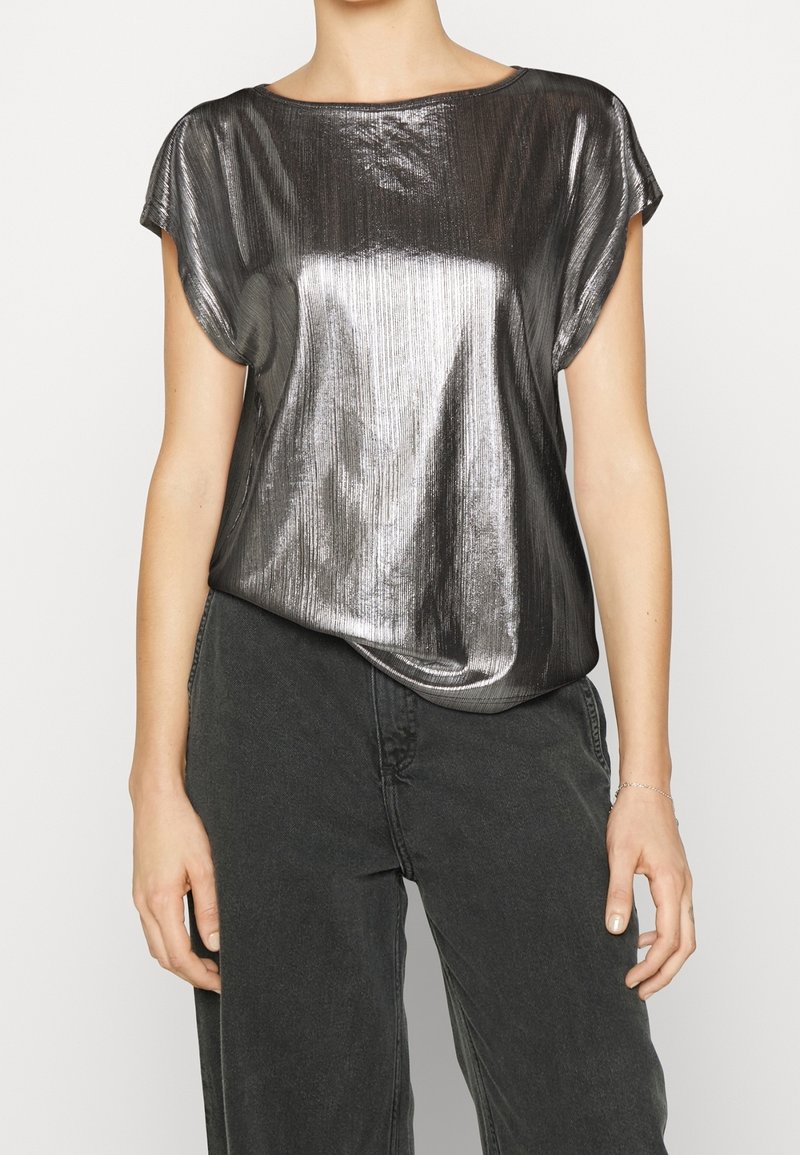 Femme portant un haut métallique brillant à manches courtes associé à un pantalon noir taille haute, montrant le haut du corps et les bras sur un fond neutre.