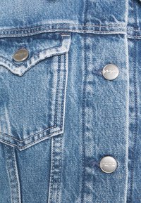 Denim jacket in light blue, featuring two chest pockets, silver metal buttons, visible stitching, and a textured fabric finish.