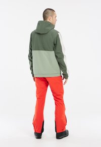 Hættejakke i grøn og lys grøn, med et farveblokdesign. Kombineret med lys orange bukser med sorte detaljer og støvler.