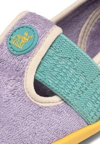 Chaussure texturée violette pour tout-petit avec bordure beige, bride réglable bleu sarcelle, semelle jaune et petit badge rond bleu sarcelle illustrant le contour d’un chien jaune.