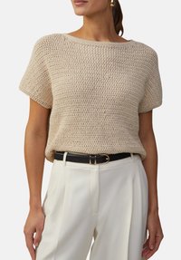 Beige gebreide top met korte mouwen, ronde halslijn en open geweven textuur, gecombineerd met hoge witte broeken en een zwarte riem.