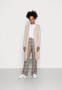 Pikk beež kardigan valge T-särgi peal, kombineeritud laia lõikega ruuduliste pükstega toonides kreem, pruun ja must, täiustatud valgete tossudega.