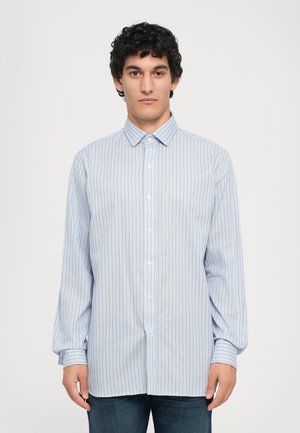 Joven con cabello rizado y oscuro, vistiendo una camisa de rayas de manga larga en azul claro y jeans oscuros, de pie contra un fondo blanco sencillo.