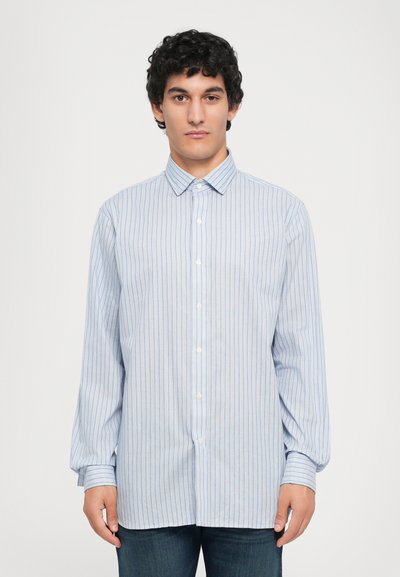 Joven con cabello rizado y oscuro, vistiendo una camisa de rayas de manga larga en azul claro y jeans oscuros, de pie contra un fondo blanco sencillo.
