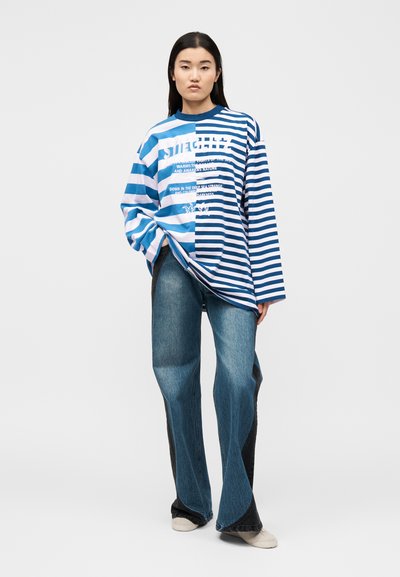 Γυναίκα που στέκεται, φοράει μπλούζα μισοριγέ σε μπλε και άσπρο με μακριά μανίκια και κείμενο, δίχρωμο φαρδύ τζιν και ανοιχτόχρωμα παπούτσια.