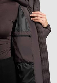 Topla, quiltana smeđa jakna s sjajnom crnom unutrašnjošću, koja ima zatvoreni džep na lijevoj strani i rebraste detalje duž rubova.