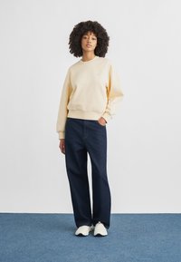 Ανοιχτό κίτρινο φούτερ με ριμπ μανσέτες, συνδυασμένο με σκούρο μπλε τζιν παντελόνι με φαρδιά πόδια και λευκά αθλητικά παπούτσια. Απλός σχεδιασμός, ελάχιστες λεπτομέρειες.