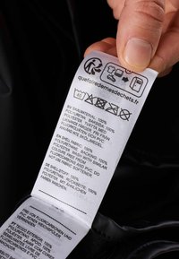 Klädselmärkning med vit tyg och svart text, instruktioner och symboler som anger 100% polyurethane och polyester material.