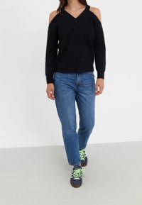 Pull noir en maille avec épaules dénudées, col en V et manches longues, porté avec un jean bleu et des baskets aux lacets vert néon.