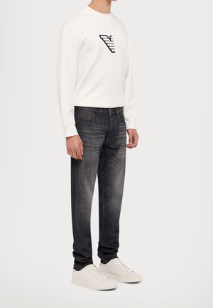 Homme portant un sweat-shirt blanc avec un logo géométrique, un jean foncé délavé et des baskets blanches, debout sur un fond clair uni.