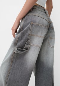 Szare, wysokie dżinsy z efektem ombre od jasnego do ciemnego, z wieloma kieszeniami i kontrastowym pomarańczowym przeszyciem. Miękka tekstura dżinsu.