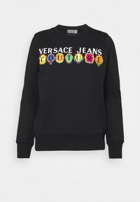 Svart hoodtröja med rund halsringning, prydd med texten "VERSACE JEANS COUTURE" i färgglada, utsmyckade bokstäver över framsidan. Mjukt tyg.