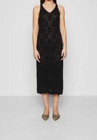 Žena v bezrukávových černých pletených midi šatech s chevronovým výřezem a bronzových špičatých balerínách, stojící na jednobarevném pozadí.