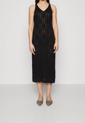 Kvinde iført en ærmeløs sort strikket midi-kjole med chevron-udskæringsmønster og bronzefarvede spidse flade sko, stående foran en ensfarvet baggrund.