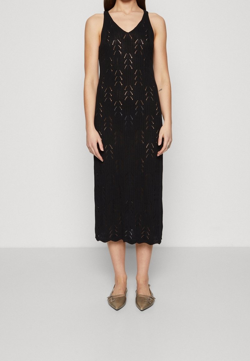 Žena v bezrukávových černých pletených midi šatech s chevronovým výřezem a bronzových špičatých balerínách, stojící na jednobarevném pozadí.